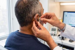 Audioprothésiste réglant une aide auditive discrète sur un patient vu de dos en cabinet